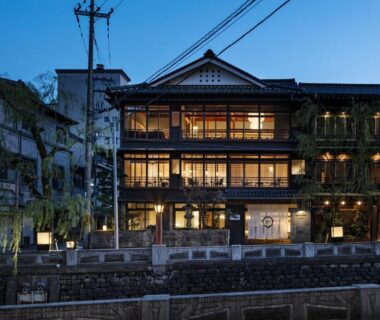 Illuminated multi-storey wooden ryokan facade along a riverside embankment in Kinosaki Onsen, Hyōgo at dusk