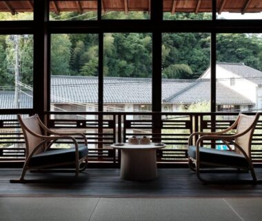 Tatami-floor seating with low table and chairs facing large windows overlooking tiled roofs and forested hillside