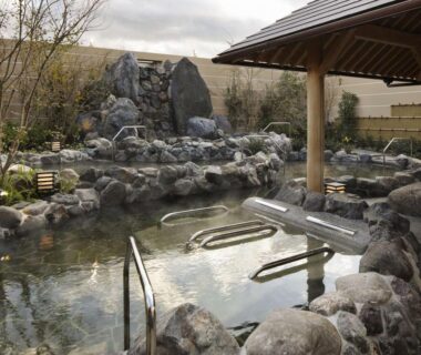 Rock-lined outdoor onsen with wooden shelter and metal handrails at Kobe Minato Onsen Ren, Hyōgo