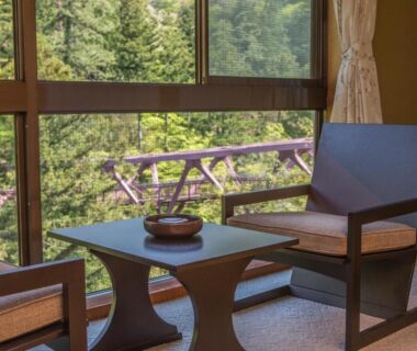 Two wooden chairs and low table by window overlooking green forest and balcony at Kocho ryokan, Ishikawa
