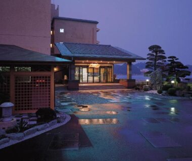 Lit outdoor stone courtyard with entrance, pine trees and distant city lights at Kochoran in Mie