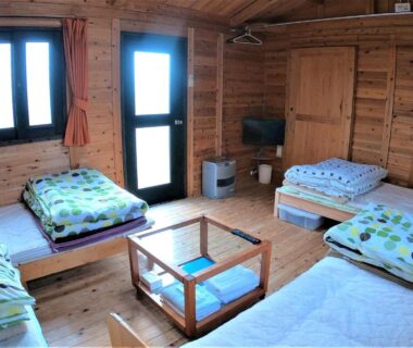 Shared wood-paneled guest room with multiple futon beds, low glass table, TV, and exterior door at Koinami, Mie