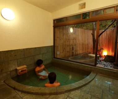 Guests soak in a circular indoor onsen tub beside a small outdoor bathing area with bamboo fence