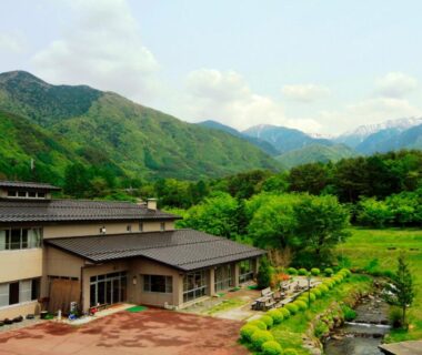Exterior of Komao ryokan with tiled roofs, garden and stream against Nagano mountain backdrop