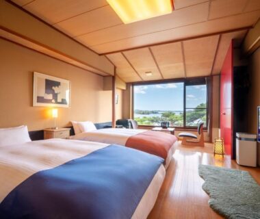 Twin beds with blue and orange runners, wooden interior, low seating and large window overlooking green landscape