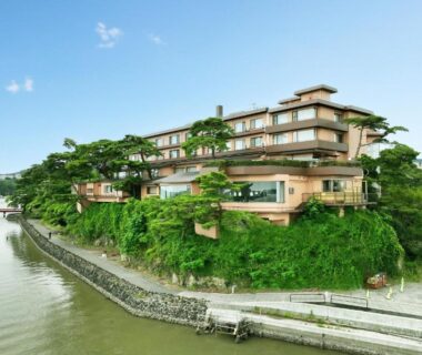 Multi-story ryokan perched on river bend with balconies, stone embankment and surrounding trees in Miyagi, Tōhoku