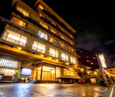 Lit multi-story ryokan exterior at night with warm lantern-lit entrance and street in Shizuoka