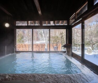 Steaming indoor onsen bath with wooden beams and floor-to-ceiling windows overlooking snow-covered garden at Konashi No Yu Sasaya, Nagano