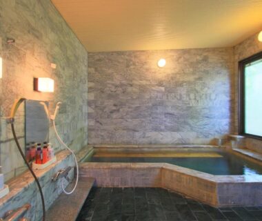 Stone-walled indoor bath with wooden-lined tub, wall-mounted showers and window at Kounso ryokan, Yamanashi