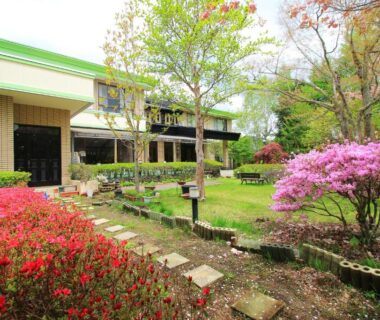 Stepping stone garden path with pink flowering shrubs and green lawn beside Kounso ryokan building in Yamanashi