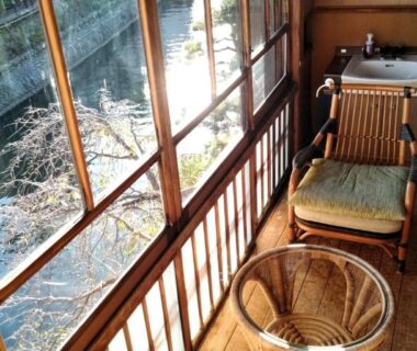 Wooden riverside alcove with wicker chair and low table overlooking a stream through large windows