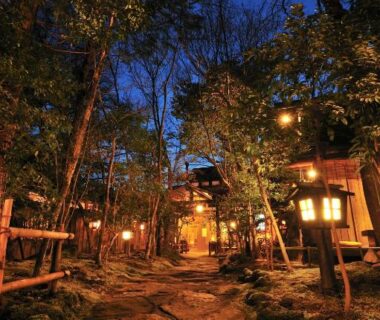 Stone pathway flanked by lanterns and trees leading to wooden ryokan buildings at dusk in Kurokawa Onsen, Kumamoto