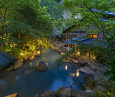 Stone-lined outdoor onsen bath flanked by wooden ryokan buildings and lanterns among lush green trees at dusk