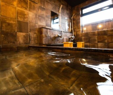 Reflective indoor onsen bath with tiled walls, wooden stools and shower area at Kurosakisou, Kyoto