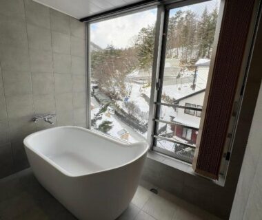 Freestanding bathtub beside floor-to-ceiling window overlooking snowy rooftops and forested hillside in Kusatsu, Gunma