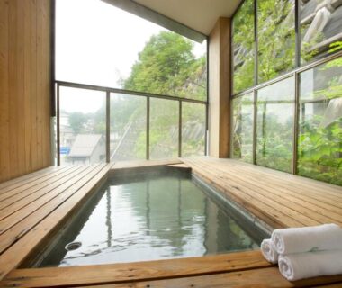 Wooden indoor bath with rolled towels by floor-to-ceiling glass and hillside greenery at Kusatsu Hotel Bekkan