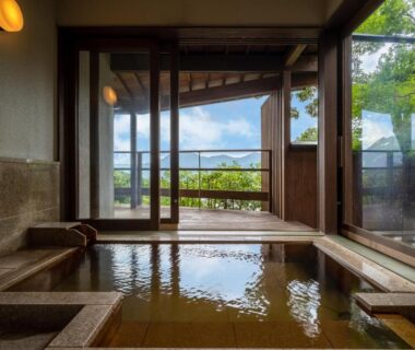 Indoor onsen bath with stone edging, sliding glass doors opening to terrace and hillside view at Ryunohige, Ōita