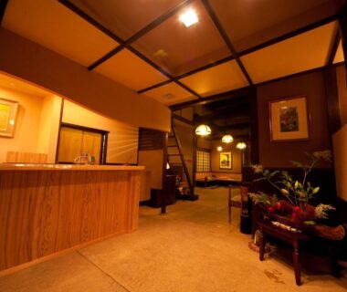 Ryokan reception with wooden counter, low-beamed ceiling, seating area, potted plants and warm lighting in Ōita