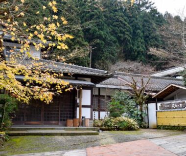 Traditional wooden ryokan entrance with sliding doors, tiled courtyard and forested hillside in Kyoto