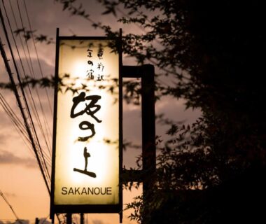 Backlit vertical sign reading SAKANOUE at dusk, framed by trees and utility lines against the sky