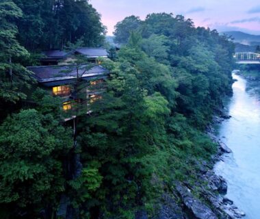 Multi-story ryokan building lit beside a forested riverbank at Kyousen in Nagano