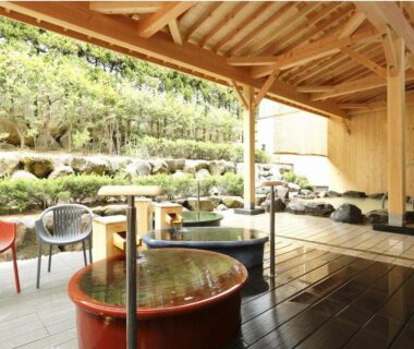 Covered wooden terrace with round soaking tubs, stone baths, chairs and landscaped garden at Kyukamura Noto-Chirihama, Ishikawa