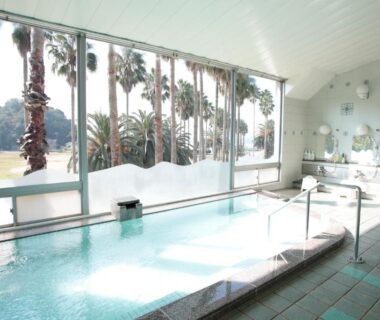 Bright indoor bathing pool with large windows overlooking palm trees at Kyukamura Ohkunoshima Hiroshima