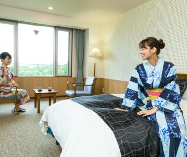 Two women in yukata relaxing in a ryokan room with bed, seating area and green view through window