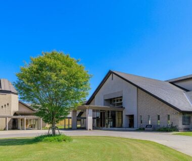 Exterior view of ryokan entrance with green lawn and lone tree under clear blue sky in Fukushima