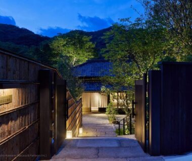 Stone steps leading through wooden gates to a lit ryokan entrance with trees and mountain backdrop at dusk