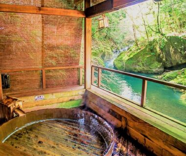 Round wooden soaking tub on open veranda overlooking emerald river and moss-covered rocks in Shizuoka
