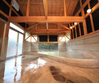 Spacious indoor onsen with wooden beams, stone floor and large windows at Lodge Toranoyu, Ōita, Kyūshū