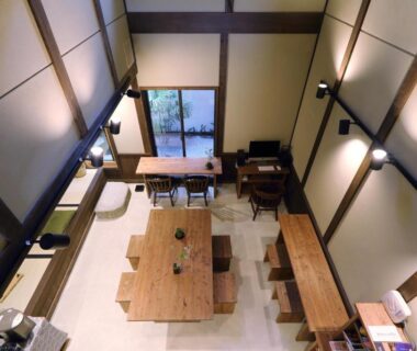 Overhead view of communal dining area with long wooden table, benches, exposed beams and window overlooking courtyard