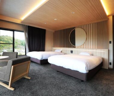 Twin beds, wooden slatted wall, round mirror, lounge chairs and large window overlooking greenery at Manten no Tsujinoya, Ishikawa