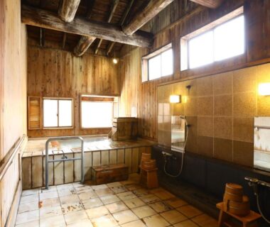 Indoor wooden onsen with tiled floor, soaking tub, washing stools and mirrors at Manza Onsen Nisshinkan in Gunma