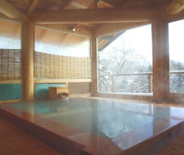 Steaming indoor onsen pool with wooden beams, tiled bath and large windows overlooking wooded hillside