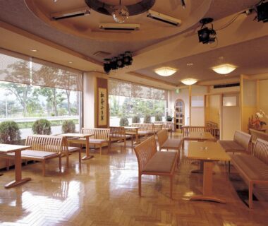 Bright lounge with wooden floors, upholstered benches and low tables by large windows overlooking trees, Maruei, Yamanashi