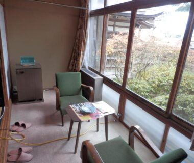 Carpeted sitting area with low table, green chairs, slippers and large windows overlooking garden at Maruka Ryokan in Nagano