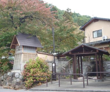 Small wooden shelter with stone steps and lantern beside a two-story ryokan on a tree-covered hill in Nagano