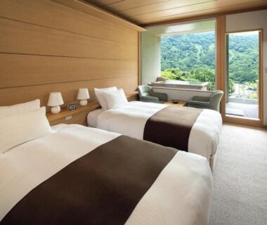 Twin beds with wooden soaking tub and seating by window overlooking green mountains