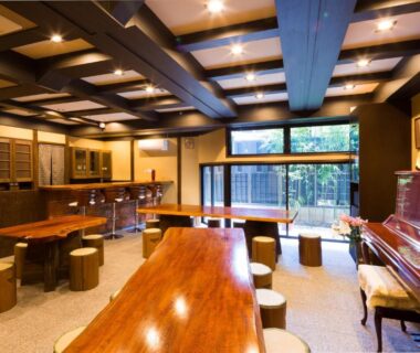 Interior lounge with wooden tables, stools, piano and large windows overlooking greenery at Matsubaya Ryokan, Kyoto