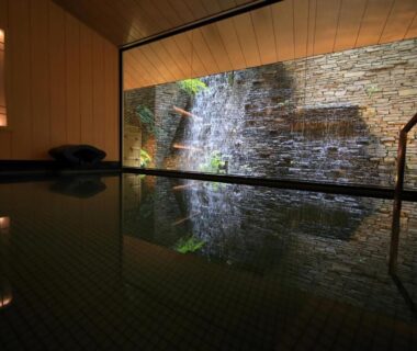 Indoor onsen bath with stone waterfall wall, wooden ceiling, reflective water surface and handrail at Matsui Honkan Kyoto