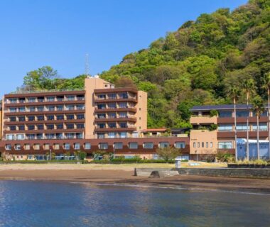 Multi-story ryokan on shoreline with balconies and forested hillside in Shizuoka, Chūbu region