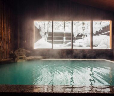Wood-lined indoor onsen bath with steamy water and snow visible through large windows at Meitoya So Yamagata