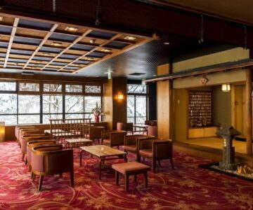 Spacious ryokan lounge with chairs, low tables, tatami alcove and large windows overlooking trees