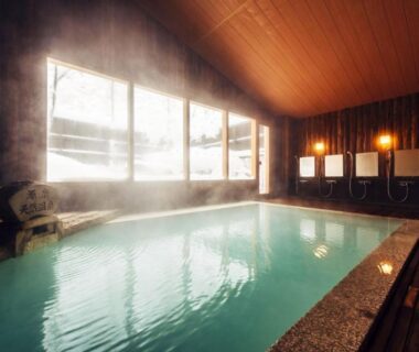 Indoor onsen bath with wooden vaulted ceiling, stone water spout, tiled pool and wash stations along the wall