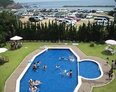 Guests in outdoor pool with lawn seating umbrellas and coastal parking visible at Michishio in Mie