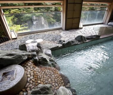 Indoor onsen bath with stone edging and windows overlooking a riverside forest at Minakami Hotel Juraku, Gunma