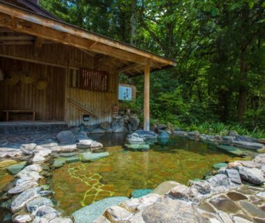 Stone-lined outdoor onsen beside a wooden shelter surrounded by forest at Minakamisansou in Gunma