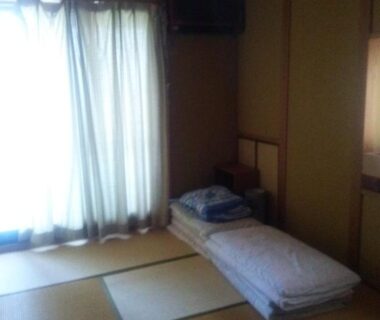 Simple tatami room with folded futon by curtained window at Minshuku Kamagari, Hiroshima
